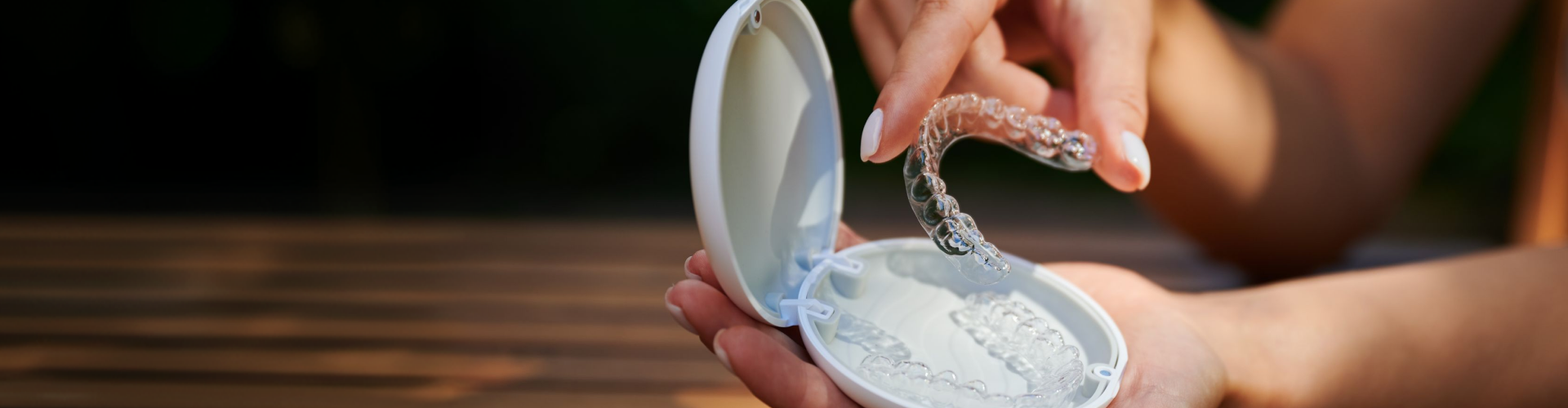 Hand holding a clear dental aligner above an open case containing additional transparent aligner trays.